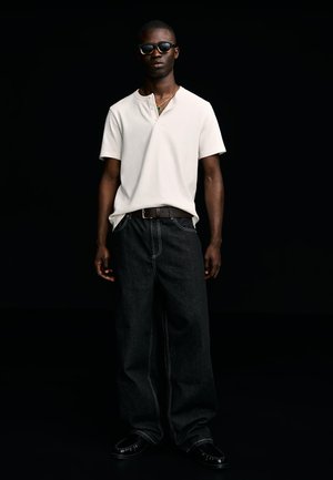 Moški z črnimi sončnimi očali, kratkimi rokavi bele henley majice, širokimi temnimi kavbojkami z belim šivom, črnim pasom in črnimi čevlji.