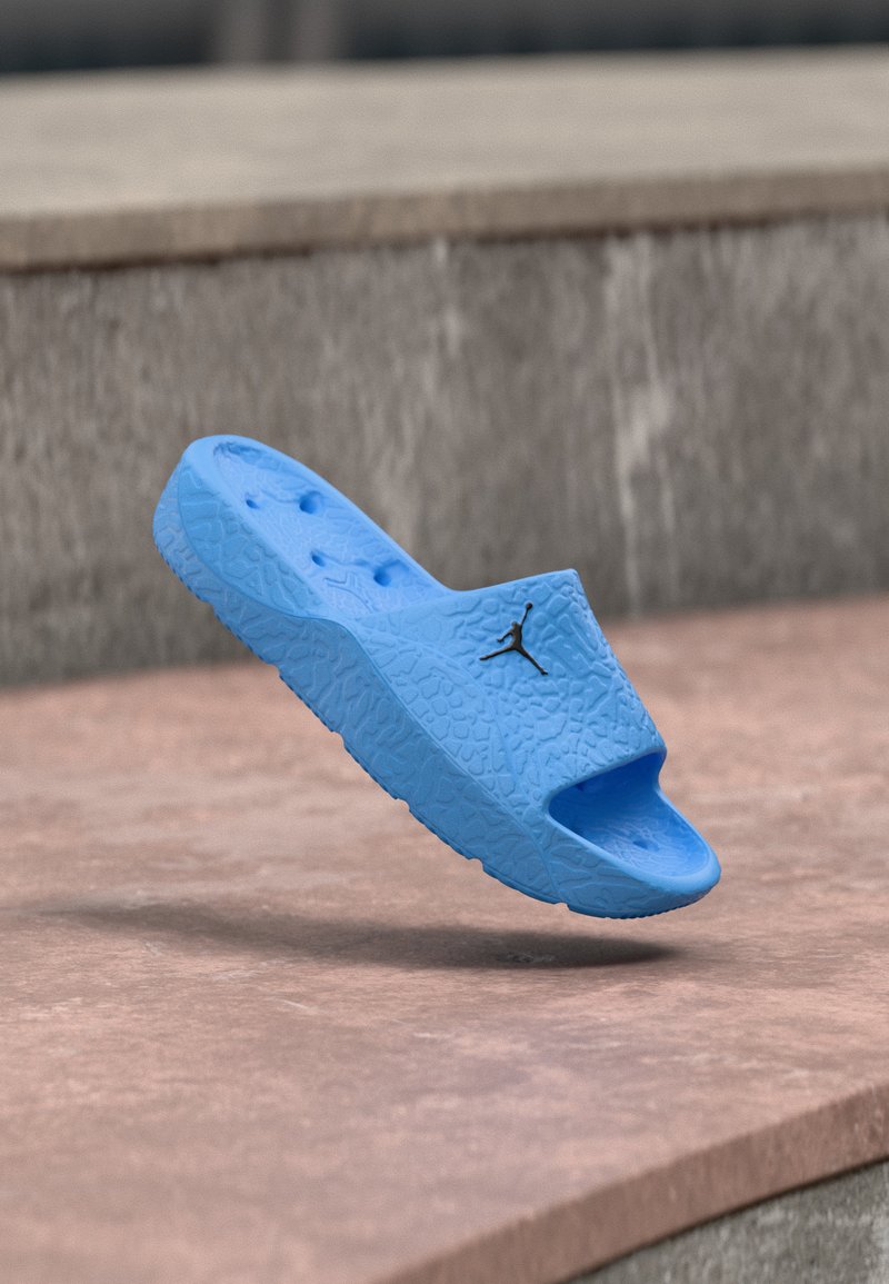 Μπλε υφασμάτινο σανδάλι slide με μαύρο λογότυπο Jumpman που αιωρείται πάνω από πέτρινα σκαλοπάτια σε εξωτερική επιφάνεια.