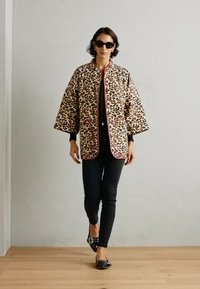 Jacke mit Leopardenmuster und pinken Akzenten, oversized Schnitt, kombiniert mit schwarzem Oberteil und schmal geschnittenen Hosen. Model trägt dunkle Sonnenbrille und Ballerinas.