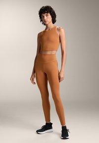 Szorosan illeszkedő, ujjatlan crop top és magas derékú leggings rozsdaszínben, sima rugalmas anyagból. Fekete sportcipők.