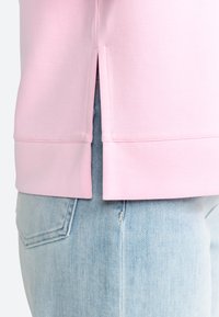Sweatshirt rose avec un détail d'herbe sur le côté, texture lisse et ourlet côtelé. Porté par-dessus un denim bleu clair, mettant en valeur un style décontracté.