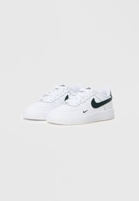 Bijele Nike niske tenisice s crnim swoosh logotipima, vezice vezane, prikazane na jednostavnoj svijetlosivoj pozadini.