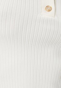 Witte ribgebreide top met een gestructureerd breipatroon, afgewerkt met een knoopdetail in goudkleurige hardware.