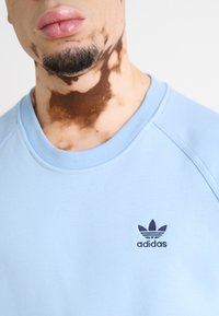 Svetlo modra majica s krojačem in raglan rokavi. Vključuje majhen črn Adidas logotip na prsih. Gladka tekstura tkanine.