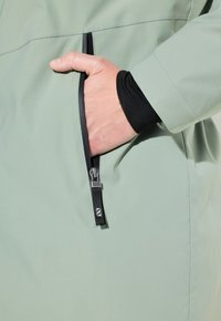 Giacca verde chiaro con tasca con zip, design elegante, texture liscia e dettaglio della cerniera nera. Mano inserita nella tasca.