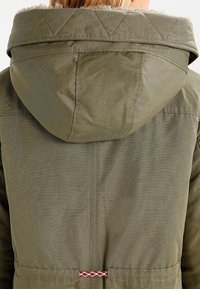 Chaqueta con capucha de color verde oliva con acentos acolchados, tejido texturizado y un cordón trenzado en la cintura. La vista desde atrás muestra costuras verticales.
