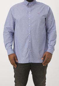 Man in een blauw en wit geruit overhemd met knopen en donkere broek, staand tegen een effen achtergrond, met handen ontspannen langs de zij.