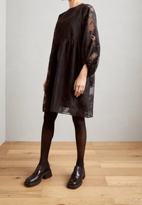Robe noire fluide avec des manches transparentes texturées et un col arrondi. Associée à des mocassins noirs et des collants opaques. Fond de sol en bois.