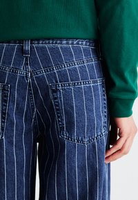 Striped blue denim jeans with white vertical lines, featuring two back pockets and a straight cut. A green top partially visible above.