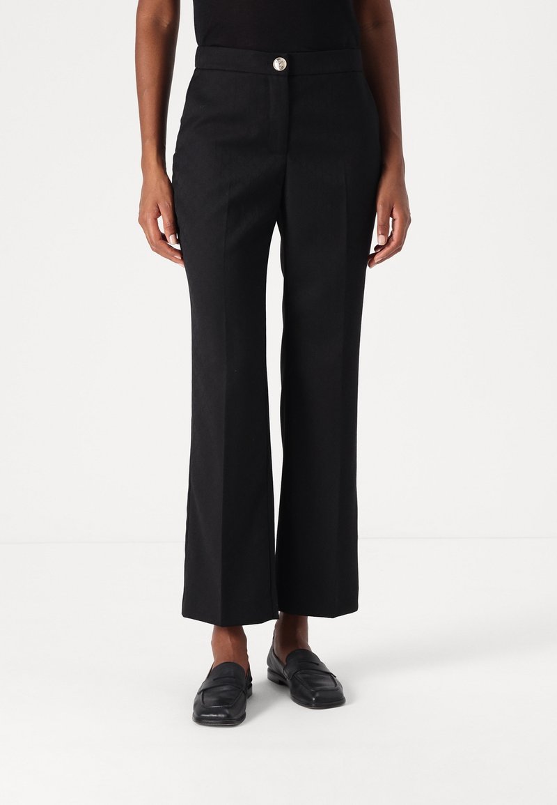 Femme portant un pantalon noir taille haute à jambes larges avec un pli frontal et des mocassins noirs, debout devant un fond blanc.