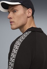 Tânăr închis la culoare cu o șapcă neagră și un tricou negru cu dungi albe pe manșon, privind spre stânga pe un fundal gri.