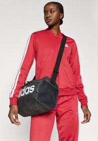 Crna Adidas torba za sport s teksturiranom površinom, bijelim logotipom i podesivim remenom. Model nosi crveni trenirku s bijelim prugama.