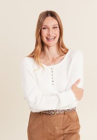 Maglione bianco di pelliccia con scollatura tonda e cinque bottoni sul davanti, abbinato a pantaloni marroni e una cintura con stampa leopardata.