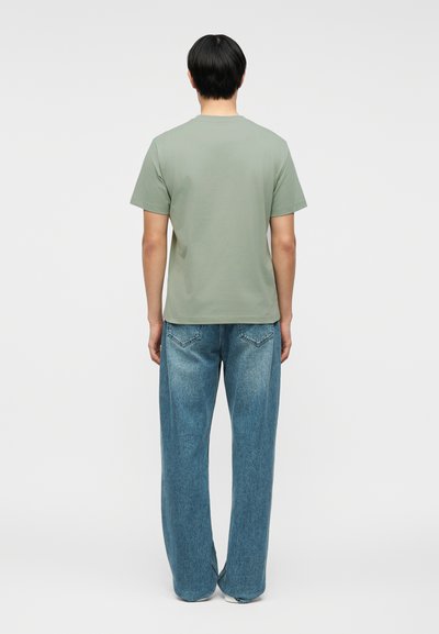 Άτομο που στέκεται γυρισμένο πλάτη, φοράει απλό πράσινο πουπουλένιο μπλουζάκι και φαρδιά μπλε τζιν, μπροστά σε απλό γκρι φόντο.