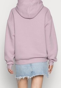 Sudadera rosa claro de gran tamaño con capucha, puños y dobladillo acanalados, combinada con una falda de mezclilla deshilachada. Textura suave, diseño casual.