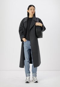 Lungo cappotto di lana grigio con ampio colletto, vestibilità comoda e maniche arrotolate, abbinato a un top nero, jeans azzurri chiari e sneaker argento.