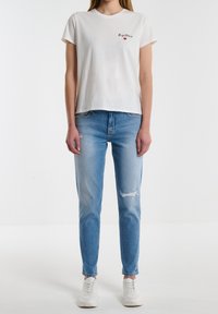 Camiseta de algodón blanca con el texto "Baster" y detalle de corazón rojo, combinada con jeans de mezclilla azul claro con una rodilla desgastada. Zapatillas blancas.