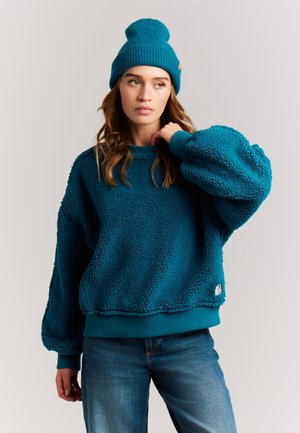 Jonge vrouw met een turquoise gebreide muts, een turquoise fleece trui met ballonmouwen en een blauwe spijkerbroek, kijkt zijwaarts met een neutrale uitdrukking.
