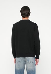 Oseba, oblečena v enostavno črno dolgesleeve kapuco in svetlo modre pošle sajne jeans, stoji obrnjena proti bel fokus.