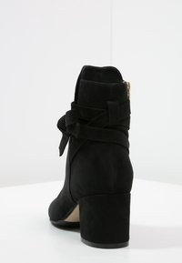 Bottine cheville en daim noir avec talon bloc, détail de sangle enroulante et fermeture éclair latérale, posée sur une surface blanche avec un fond uni.