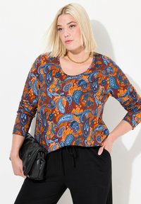 Langärmliges Oberteil mit Paisleymuster in Orange, Blau und Braun. Verfügt über einen runden Ausschnitt und einen lockeren Schnitt. Kombiniert mit schwarzen Hosen.
