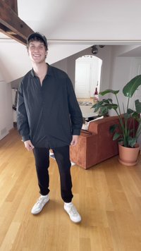 Schwarze Hemd mit Knöpfen, schwarze Hose und weiße Sneaker. Lässiger Schnitt mit langen Ärmeln. Der Hintergrund zeigt ein Sofa und Zimmerpflanzen.