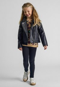 Giacca nera in ecopelle con dettagli in metallo argentato, indossata sopra un top beige con stampa leopardata colorata, abbinata a leggings scuri e sneakers bianche.