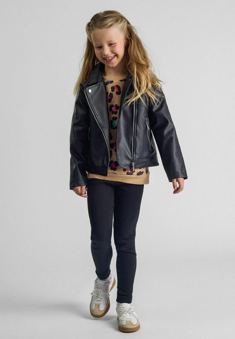Giacca nera in ecopelle con dettagli in metallo argentato, indossata sopra un top beige con stampa leopardata colorata, abbinata a leggings scuri e sneakers bianche.
