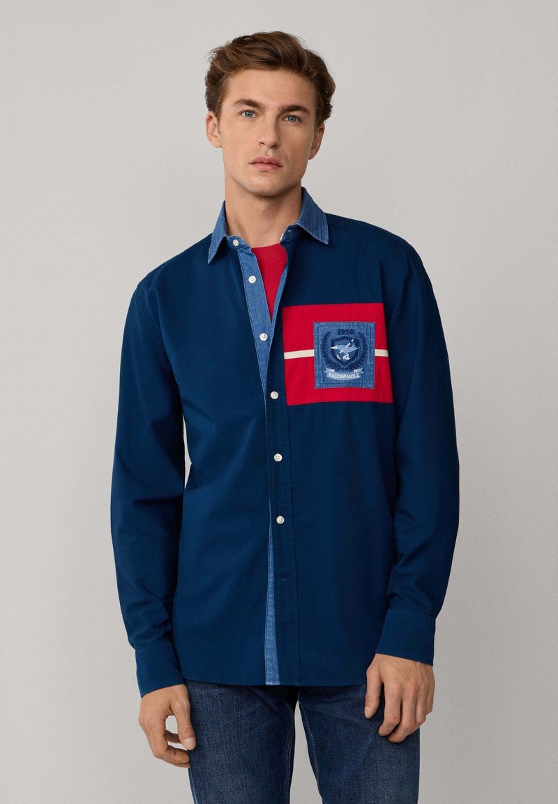 Camicia a maniche lunghe blu navy con colletto in denim e taschino sul petto con dettagli rossi e un patch con logo. Texture liscia, design con bottoni.