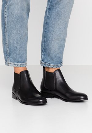Sorte læder Chelsea-støvler båret med lyseblå cropped jeans på hvidt gulv og baggrund.