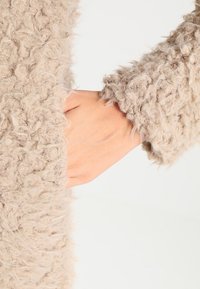 Main dans la poche d'un vêtement à manches longues beige, texturé et duveteux sur un fond clair.