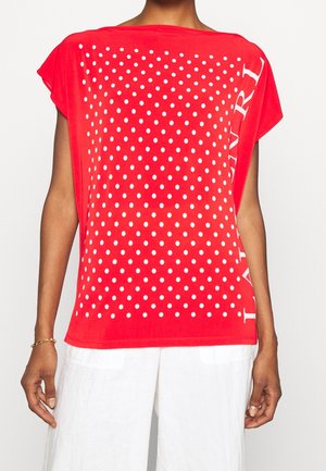 Czerwona luźna bluzka w białe grochy z częściowym pionowym białym napisem po boku, noszona z białymi spodniami.