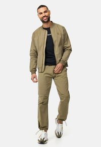Khaki bomber jacket with a zip front, black t-shirt underneath, and matching trousers. White and gray sneakers with yellow accents.