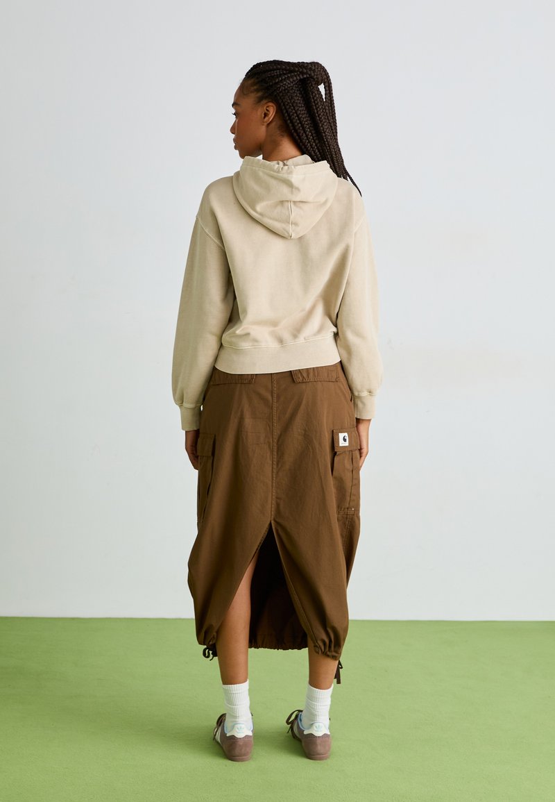 Hoodie beige avec cordon de serrage, porté par-dessus une jupe marron avec des poches latérales et une fente, associé à des chaussettes blanches et des chaussures marron.