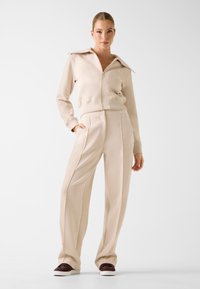 Veste beige zippée avec grand col et pantalon beige ajusté, tous deux en tissu doux à la texture lisse. Détails avec poches et boutons.