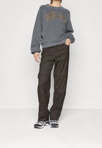 En person iklädd en grå sweatshirt med "Johnny Jeans" i leopardmönster, tillsammans med bruna byxor och sportiga skor, står mot en enfärgad bakgrund.