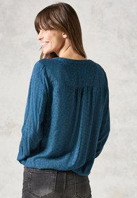 Blå långärmad topp med strukturerat mönster, rundad halsringning och veckdetalj på baksidan, kombinerad med mörka jeans.