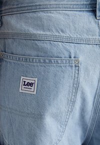 Jasnoniebieska tkanina dżinsowa z tylną kieszenią; na kieszeni znajduje się biała metka z niebieskim napisem "Lee WORKWEAR".