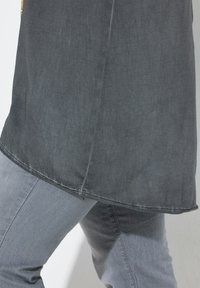 Tunique grise en tissu texturé et léger, présentant un ourlet légèrement arrondi et une fente sur le côté pour plus de liberté de mouvement, associée à un jean gris.