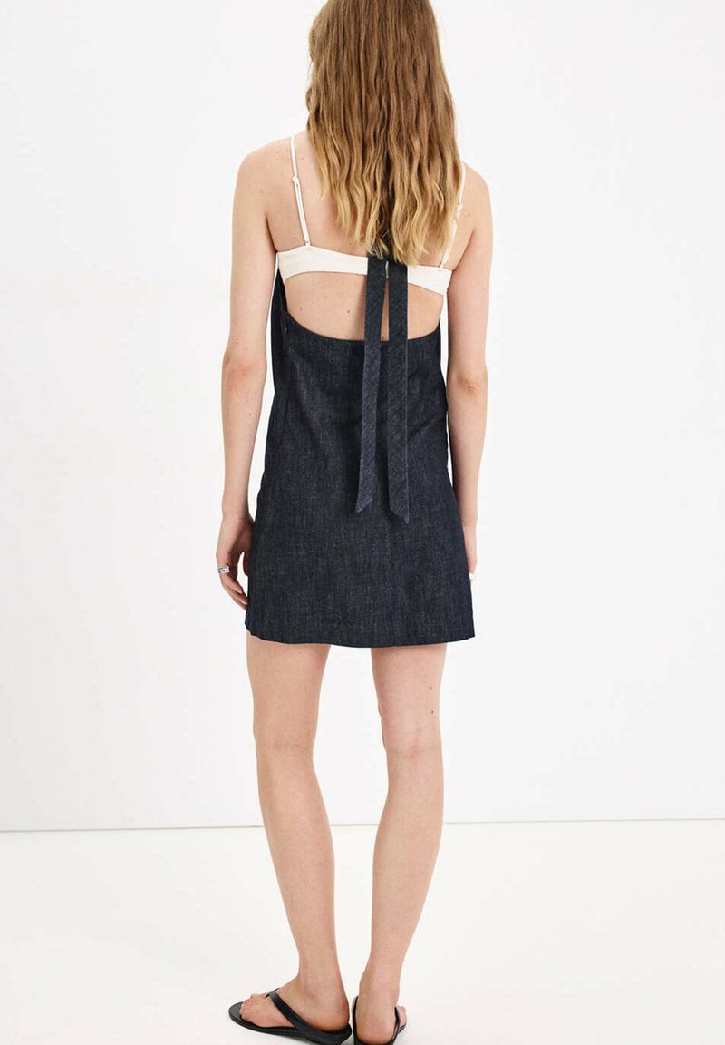 Femme aux longs cheveux porte une robe en denim sombre sans manches avec un dos ouvert et un soutien-gorge blanc, debout pieds nus sur un fond blanc.