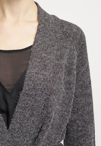 Harmaa neulecardigan, jossa on huivikaulus, pehmeä rakenne, avoin etuosa ja joka on kerroksittain mustan läpinäkyvän topin päällä, jossa on V-päätös.