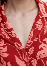 Camicia rossa con motivo floreale e colletto; presenta grandi fiori rosa e foglie; tessuto testurizzato con dettagli di cucitura visibili.