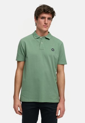 Polo en algodón verde apagado, con un collar clásico, tapeta de tres botones y un pequeño logotipo azul marino en el pecho. Mangas cortas, diseño sólido.