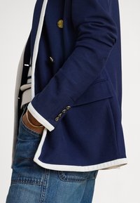 Pessoa vestindo um blazer azul-marinho com acabamento branco e botões dourados por cima de um suéter às riscas, com uma mão no bolso das calças de ganga azuis.