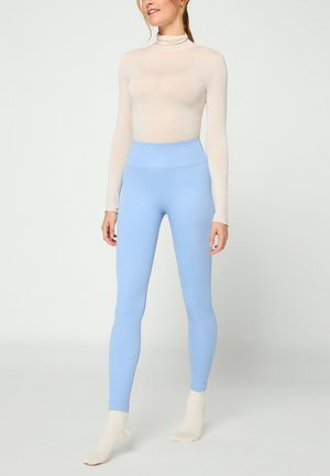 Frau steht und trägt ein beiges, langärmeliges Rollkragentop, hellblaue, hochgeschnittene Leggings und beigefarbene Socken vor einfarbigem Hintergrund.