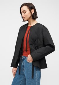 Νεαρή γυναίκα με κοντά σκούρα μαλλιά, που φοράει μια μαύρη καπιτονέ ζακέτα, μπλουζάκι σε σκουριά απόχρωση και μπλε τζιν, στέκεται έχοντας το ένα χέρι στην τσέπη.