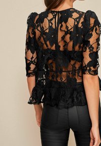 Top en dentelle noire avec un motif floral, des manches courtes bouffantes et un ourlet peplum. Le dos présente une ouverture en keyhole et un design texturé.