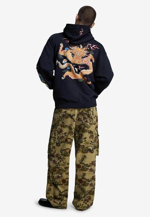 Persoon die een zwarte hoodie draagt met een grote geborduurde gouden draak op de rug en brede olijfgroene cargobroeken met bloemmotieven, staand met handen in de zakken.
