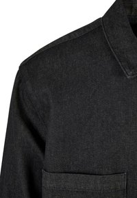 Gros plan d'une veste en jean noire montrant le col, la couture de l'épaule et le haut de la poche avant.