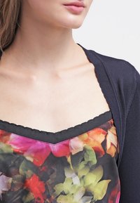 Cardigan blu navy con una finitura testurizzata indossato sopra un top a stampa floreale colorata con un collo nero ondulato.